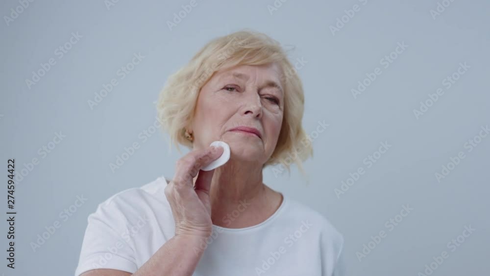Beautiful senior model cleaning her face for posing in the photo studio. Portrait of lovely blonde mature woman using a cotton disc clearing her beauty face looking in the mirror. Beautiful senior model cleaning her face for posing in the photo studio. Portrait of lovely blonde mature woman using a cotton disc clearing her beauty face looking in the mirror.