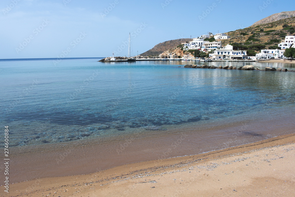 Beach at the port of Sikinos, in Greece.  View of the bay with the jetty, and village in the distance. The white buildings of the port are reflected on the calm waters.