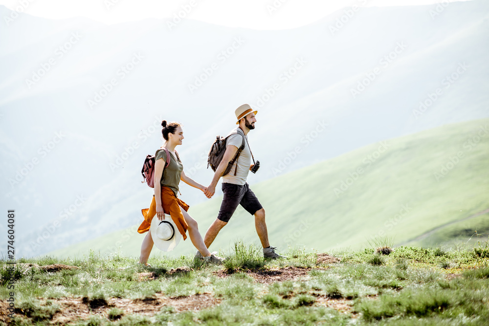 Beautiful couple walking with backpacks on the green meadow, while traveling in the mountains during the summer time - obrazy, fototapety, plakaty 