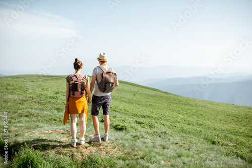 Young couple standing together with backpacks, enjoying beautiful landscape view on the green mountains, rear view