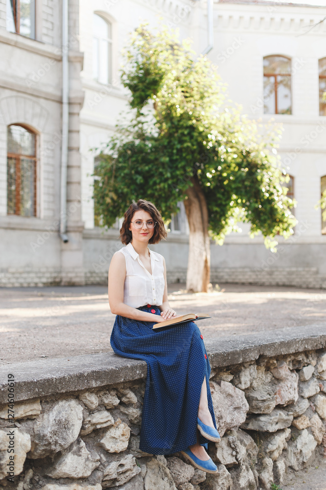 Beautiful young brunette woman with short wavy hair, wearing white sleeveless shirt, long blue skirt and glasses reading a book outdoor on old city street. Retro outfit.