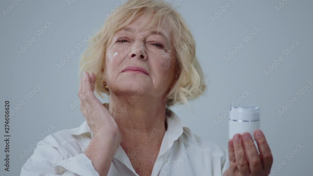 Beauty portrait of pretty senior blonde woman applying an anti-aging cream on her clear face. Close-up of mature model in white shirt holding a cream jar on gray background.