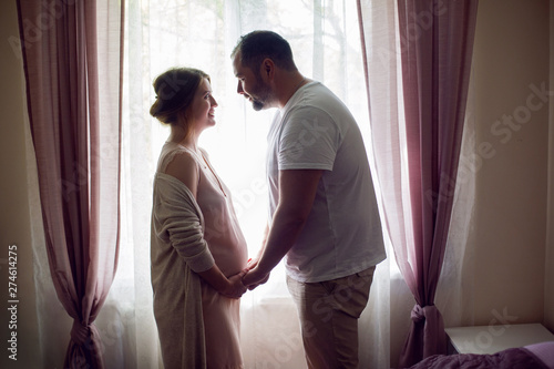 pregnant girl in a nightie with her husband in a white t-shirt stands against the window of the house