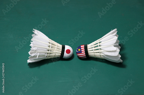 Two used shuttlecocks on green floor of Badminton court with both head each other. One head painted with Japan flag and one head painted with the Malaysia flag.