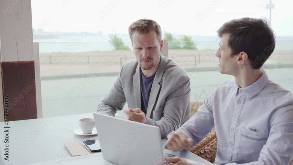 Tracking right shot of two businessmen sitting at table in cafe, looking at laptop screen and planning project. Middle aged man explaining his opinion Tracking right shot of two businessmen sitting at table in cafe, looking at laptop screen and planning project. Middle aged man explaining his opinion