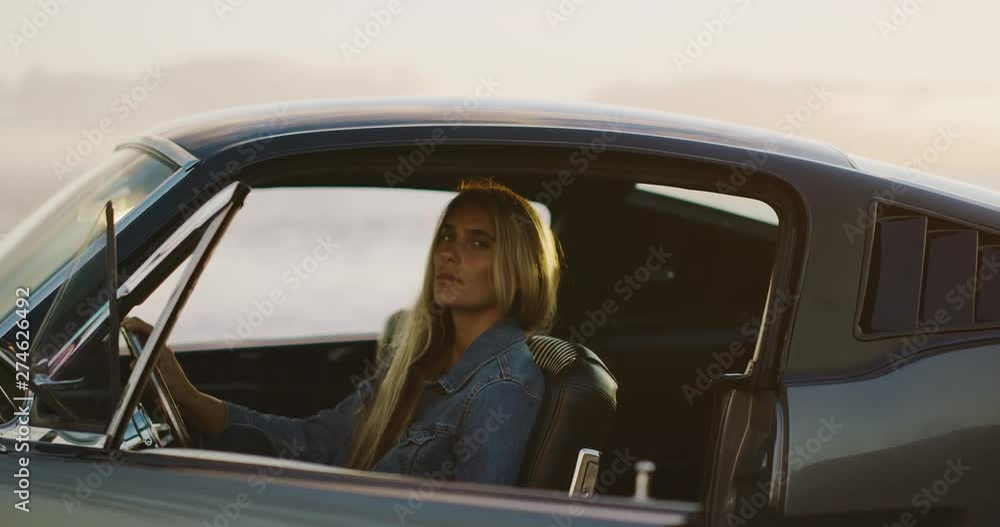 Attractive woman sitting in the driver's seat of a classic vintage sports car at sunset looking into the camera, model posing in a sports car, woman going on a solo road trip along the coast Attractive woman sitting in the driver's seat of a classic vintage sports car at sunset looking into the camera, model posing in a sports car, woman going on a solo road trip along the coast
