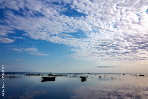 Small fishing boats at anchor, reflected in the calm and clear water of the lake, are covered with sedge in the early morning against the beautiful dawn sky of gentle pastel tones.