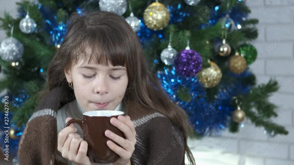 Drinking to keep warm. A cute girl wrapped in a blanket under a Christmas tree is drinking hot tea. Drinking to keep warm. A cute girl wrapped in a blanket under a Christmas tree is drinking hot tea.