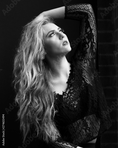 Dramatic natural light black and white portrait of a beautiful young blond woman in a lace top and bralerre with her arm raised and looking upward with a serious experssion.