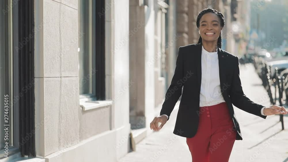 Young happy African American businesswoman in earphones walking the city street while listening to the music on the smartphone, funny dancing and looking at the camera. Outdoor. Young happy African American businesswoman in earphones walking the city street while listening to the music on the smartphone, funny dancing and looking at the camera. Outdoor.