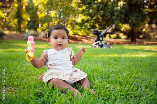 A happy hispanic baby playing and grasping a few chewable baby toys while sitting on the lawn of a park with he stroller in the background.
