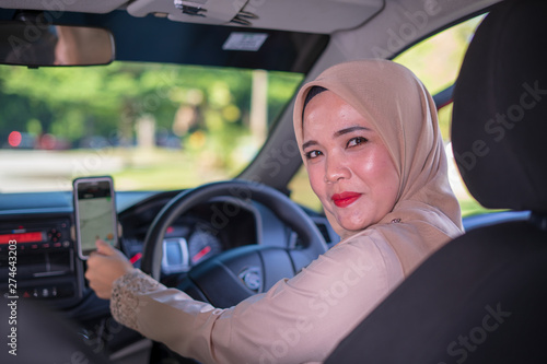 A happy young Asian Muslim woman using navigation maps on smartphone to other destination, ride sharing, preparation before driving, female driver transportation concept.