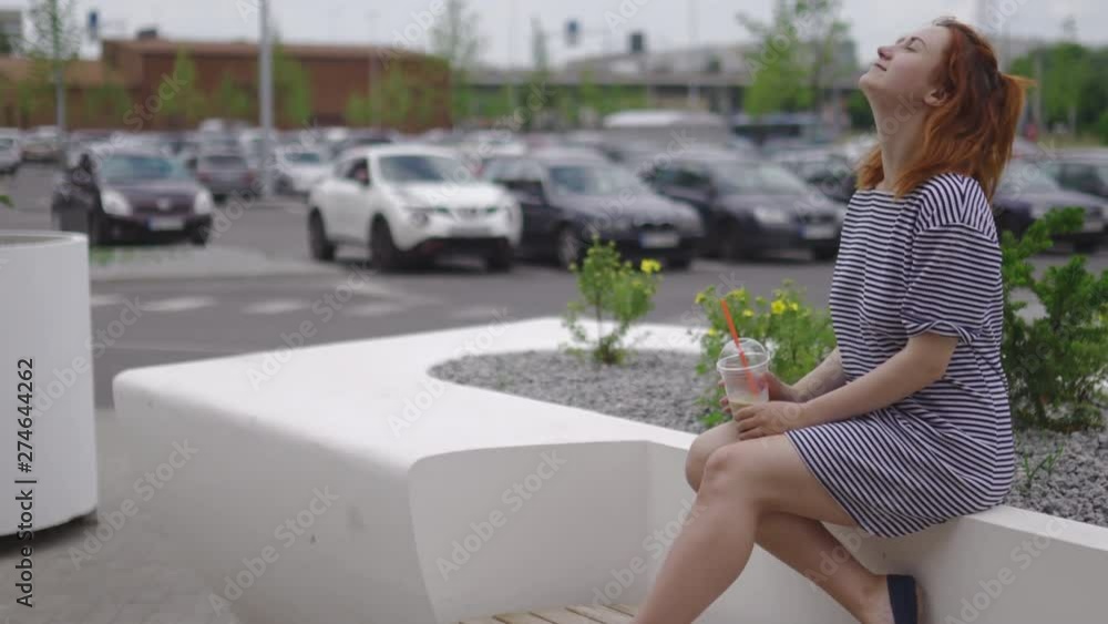 Happy young redhead woman sitting and drinking take away cold coffee wearing light striped dress in summer with cars and green leaves in the background Happy young redhead woman sitting and drinking take away cold coffee wearing light striped dress in summer with cars and green leaves in the background