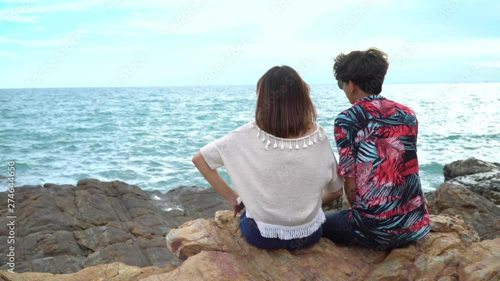 4K Medium shot rear view of young adult asian couple men and women travel tourist sitting on tropical island rocky sea coast in summer holiday vacation, they're smiling, laughing and talking together 4K Medium shot rear view of young adult asian couple men and women travel tourist sitting on tropical island rocky sea coast in summer holiday vacation, they're smiling, laughing and talking together