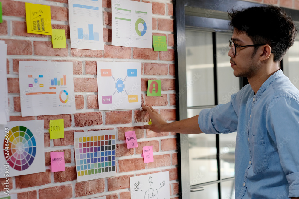 Young asian creative man reading and thinking about paper work ideas on office wall with concentration, working occupation lifestyle Young asian creative man reading and thinking about paper work ideas on office wall with concentration, working occupation lifestyle