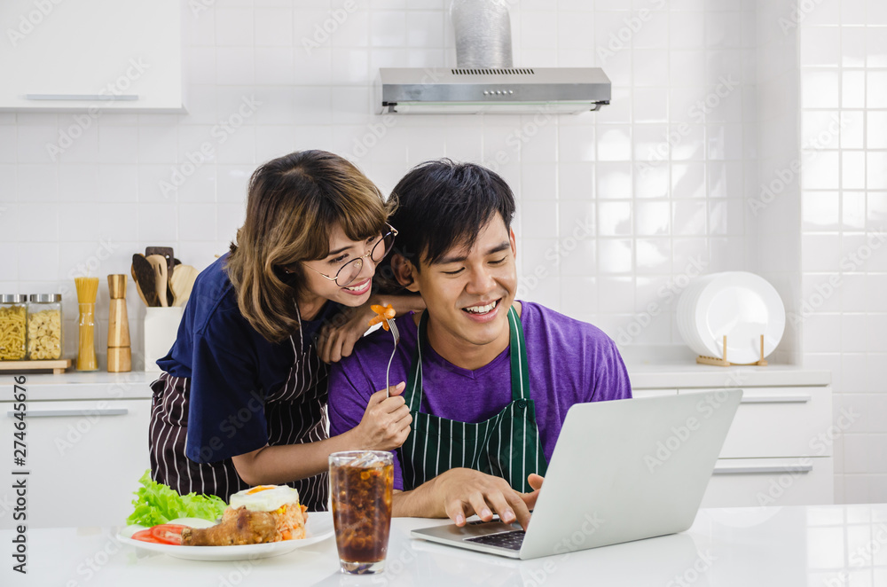 Smiling happy young Asian couple wearing apron and cooking together and feeding boyfriend while he is typing on laptop other in a white kitchen background. Smiling happy young Asian couple wearing apron and cooking together and feeding boyfriend while he is typing on laptop other in a white kitchen background.