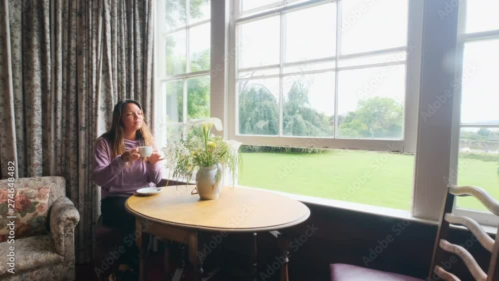 Slow motion dolly style wide daylight shot of a young woman enjoying a cup of drink at a coffee table by the window Slow motion dolly style wide daylight shot of a young woman enjoying a cup of drink at a coffee table by the window