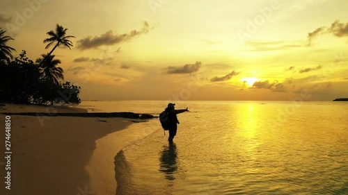 dolly shot of male tourist with backpack standing in the sea water on sandy beach and stretching hands on sunset photo