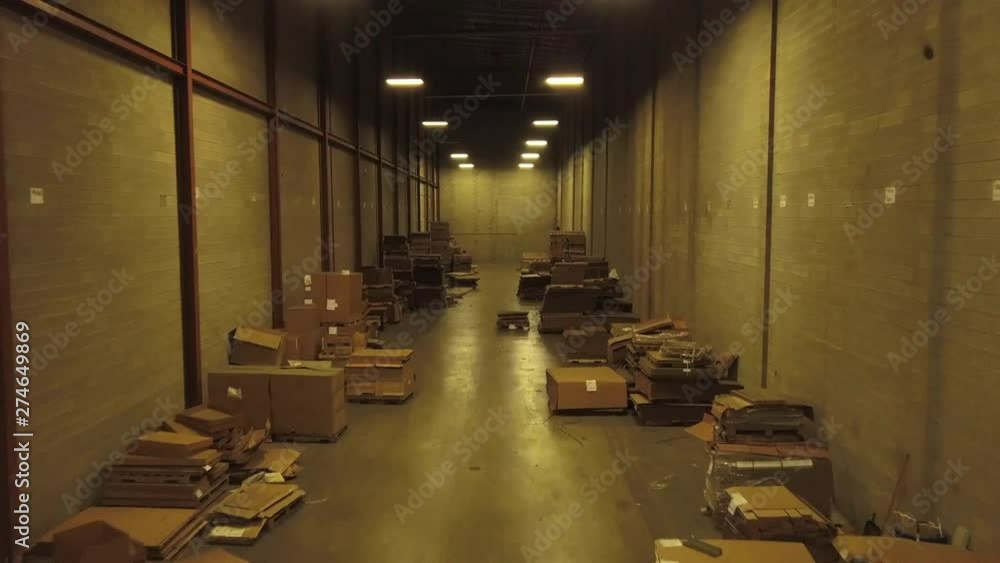 Aerial footage of boxes and files stacked on floor of a warehouse. Drone moves slowly forward between two wooden walls lined by piles of stuff on the floor.