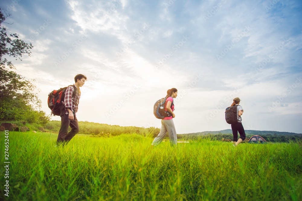 Hikers with backpacks walking through a meadow with lush grass under blue sky and sunset. Hikers with backpacks walking through a meadow with lush grass under blue sky and sunset.