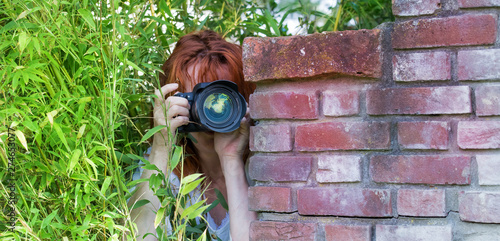 A curious woman is standing in a garden between plants and secretly observes. She takes a picture with a camera. Concept curious neighbors.