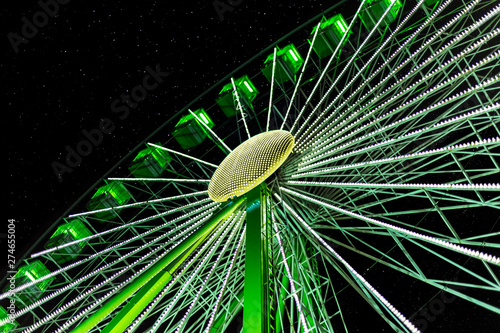 A Ferris wheel turns in the evening in the dark under a starry sky. The wheel is illuminated green and photographed diagonally from below against the sky.