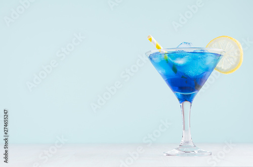 Exquisite blue cocktail for celebration in beach style with blue curacao, ice cube, lemon slice, yellow straw in mint color bar interior on white wood table. photo