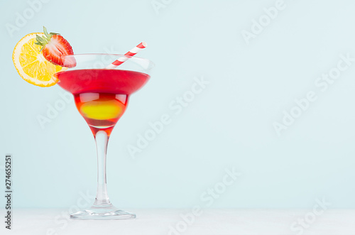 Fresh bright alcohol drink in elegant wineglass with red and yellow liquor, orange slice, strawberry, straw in modern pastel mint interior.