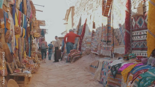 Panning through a narrow street filled with market stalls displaying colorful scarfs bags rugs and other items in an outdoor market place in a Marrakech Souk in Morocco on a sunny day