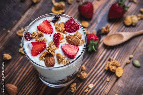 Top view bowl of homemade granola and yogurt mixed with fresh strawberries on a wooden table along with almonds on the side.