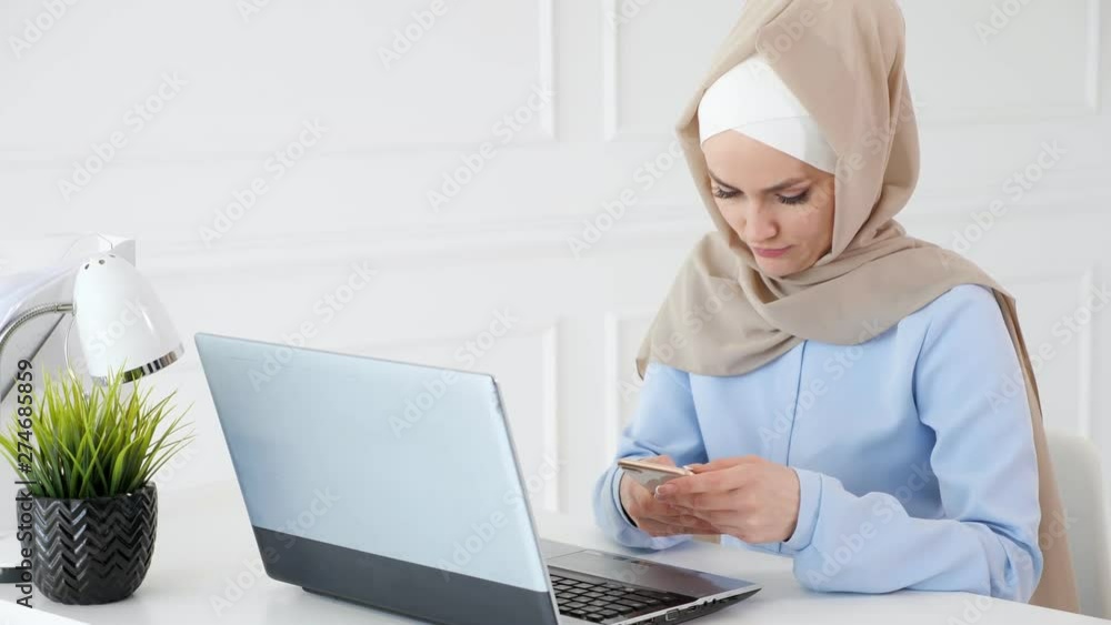 Portrait of muslim woman in beige hijab and blue dress is reading something on mobile phone sitting on her working place at table with laptop. Portrait of muslim woman in beige hijab and blue dress is reading something on mobile phone sitting on her working place at table with laptop.