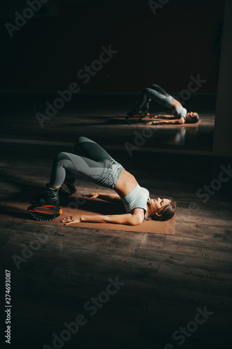 Pretty Caucasian fit brunette lying down on the mat and stretching while wearing kangoo jumps footwear. Dark gym interior.