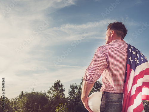 Bild auf Leinwand Young engineer, holding white hardhat and an American Flag in the park against the backdrop of green trees and the setting sun, looking into the distance
