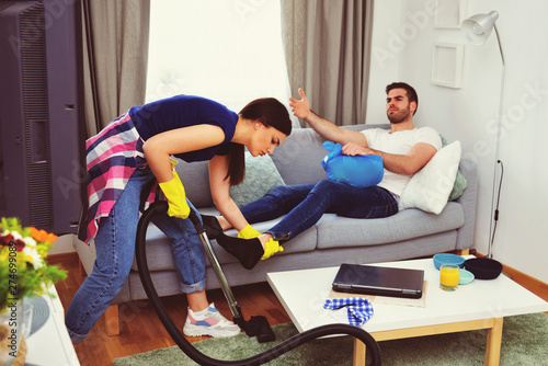 Fototapeta Naklejka Na Ścianę i Meble -  couple relationship concept, marriage  living problems and conflict, young couple in living room woman cleaning and vacuuming house, man sitting on sofa watching television, home jobs gender equality 