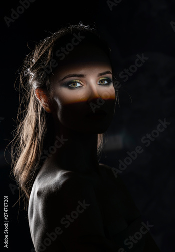 Beautiful girl model with red lips make up and naked shoulders in the shade, with a lit silhouette and a strip of light illuminating her eyes. Conceptual, art, fashionable design. Black background
