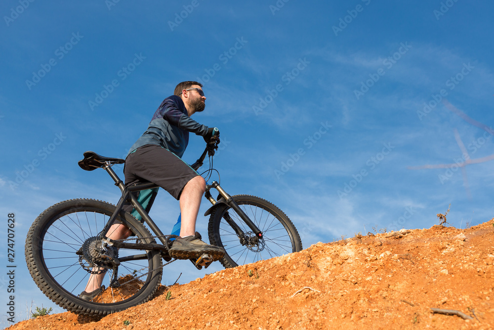 Obraz premium Cyclist in shorts and jersey on a modern carbon hardtail bike with an air suspension fork rides off-road on the orange-red hills at sunset evening in summer 