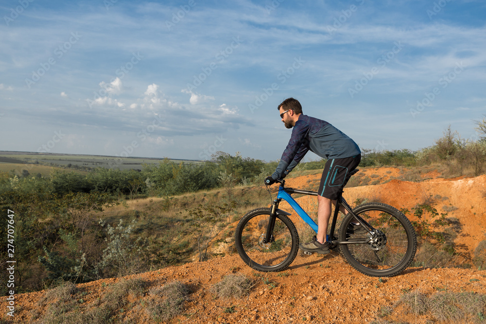 Cyclist in shorts and jersey on a modern carbon hardtail bike with an air suspension fork rides off-road on the orange-red hills at sunset evening in summer Cyclist in shorts and jersey on a modern carbon hardtail bike with an air suspension fork rides off-road on the orange-red hills at sunset evening in summer