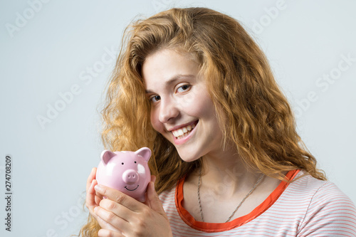 Young red-haired curly girl over white background holding piggy bank and rejoices gesturing. The concept of reliability of cash investments and insurance.