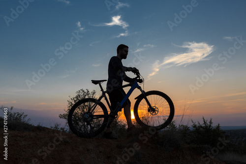 Fototapeta Naklejka Na Ścianę i Meble -  Cyclist in shorts and jersey on a modern carbon hardtail bike with an air suspension fork rides off-road on the orange-red hills at sunset evening in summer	