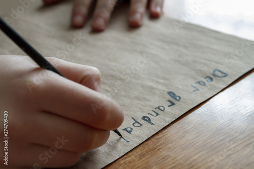A small girl writes a letter to dear grandpa by black paint on brown paper. Closeup, selective focus