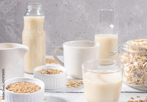 Close-up set of glass containers with oat milk, oat seeds and flakes on white table on concrete background. 