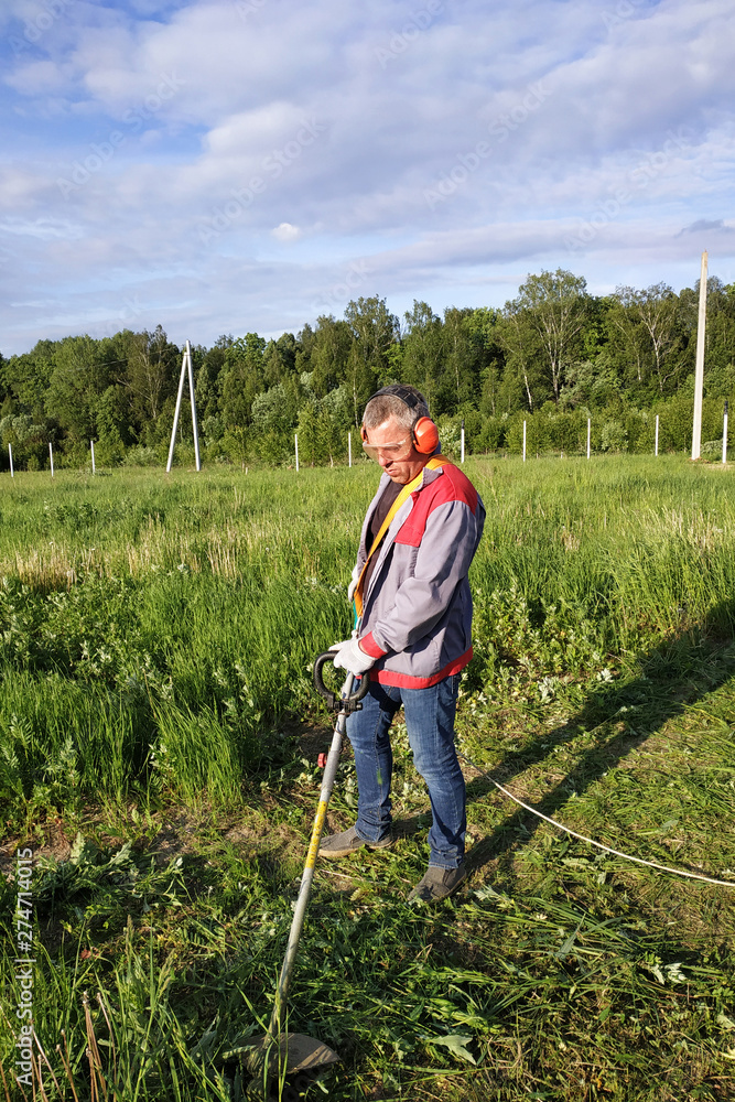 The headphone and glasses worker mows the grass in the field. The headphone and glasses worker mows the grass in the field.