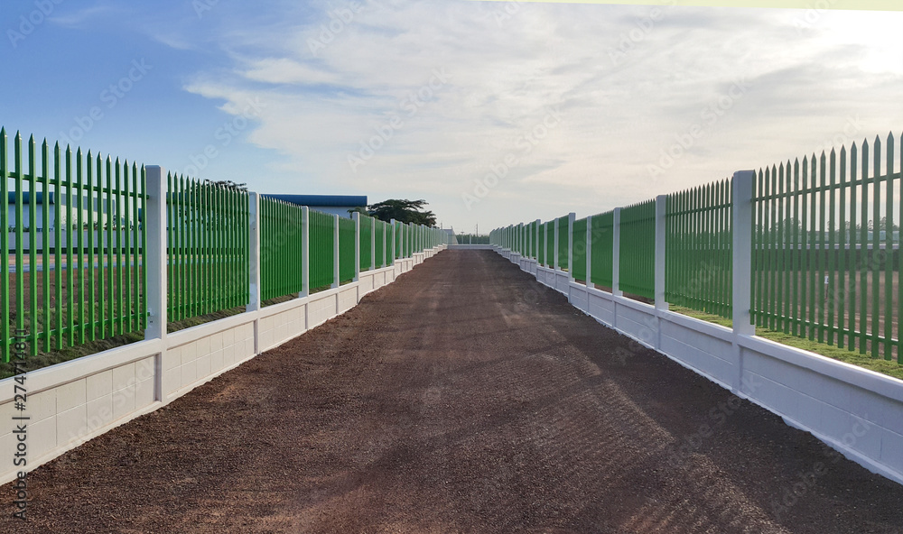 Fototapeta premium The gravel road lies between the white cement fence with green iron bars under the blue sky.