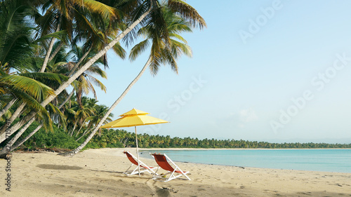 Deserted sea beach with white sand and beautiful green palm trees. Calm waves of the blue sea off the coast of Cuba. Rest on cozy red beach chairs under a yellow beach umbrella. Tropical paradise.