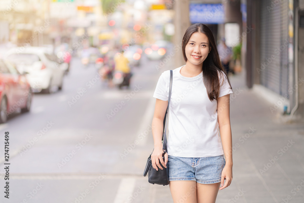 Portrait of Happy Asian young adult woman wearing white casual, short blue jeans smiling with teeth and walking on city street at sunset time. Bangkok Thailand.