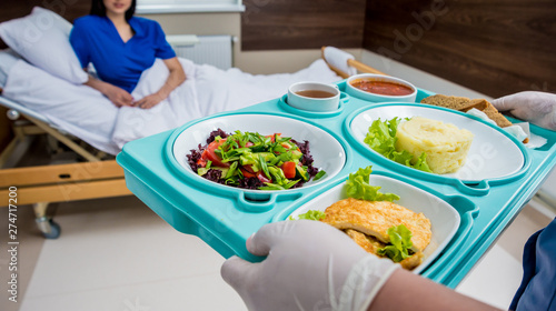 Nurse in medical coat is holding a tray with breakfast for the young female patient. © romaset