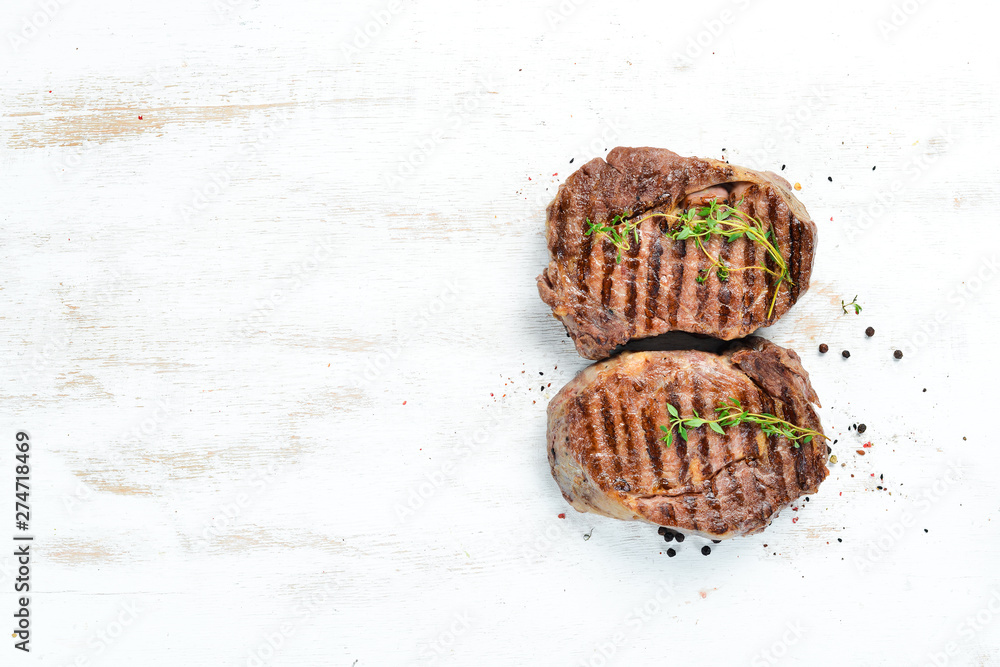 Grilled ribeye beef steak, herbs and spices on a white wooden background. Top view. Free space for your text. Grilled ribeye beef steak, herbs and spices on a white wooden background. Top view. Free space for your text.