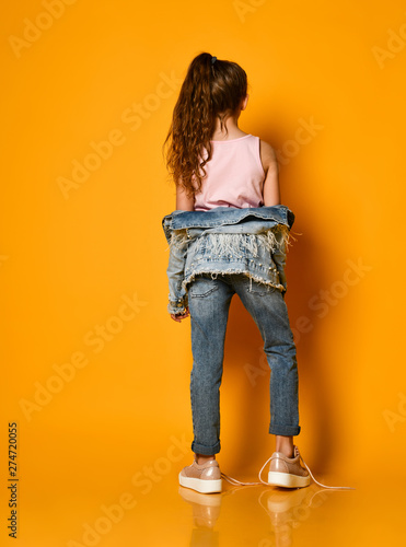 young teen girl model posing on a yellow background in jeans and a jacket, with long hair gathered in a high tail, stands half a turn to the camera. photo