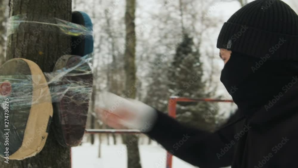 A young kickboxer guy with bandages on his hands, in a black suit trains in the fresh air in winter. He made a mannequin in a tree A young kickboxer guy with bandages on his hands, in a black suit trains in the fresh air in winter. He made a mannequin in a tree