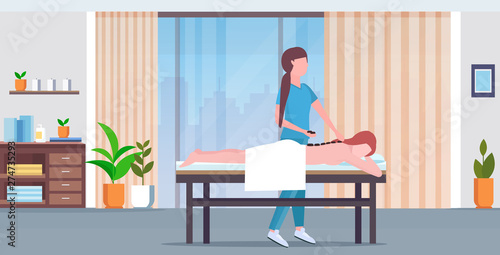 girl having hot stone back massage masseuse in uniform massaging patient body woman lying on bed treatments concept luxury spa salon clinic cabinet interior full length horizontal